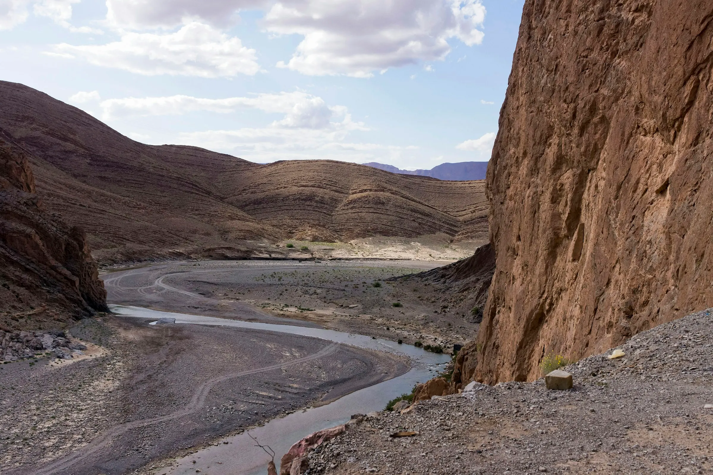 A river winding through a dry gorge of ochre, layered rock in Morocco’s Atlas Mountains under broken cloud.