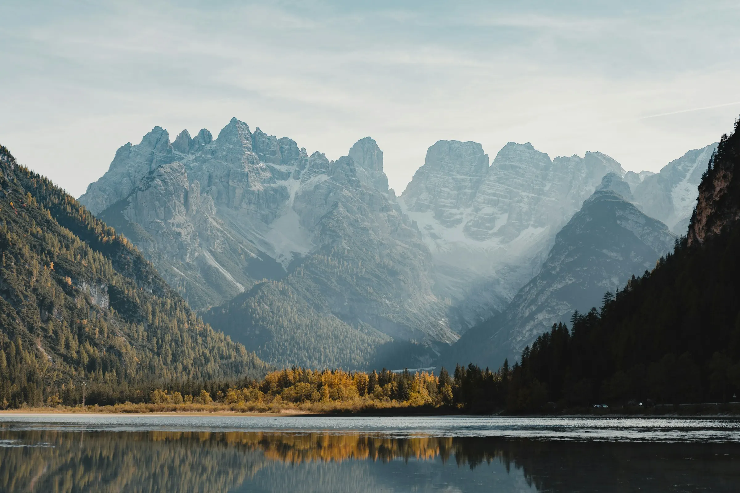 Pale limestone peaks of the Dolomites reflected in a still lake, with larches turning gold along the shore in soft autumn light.