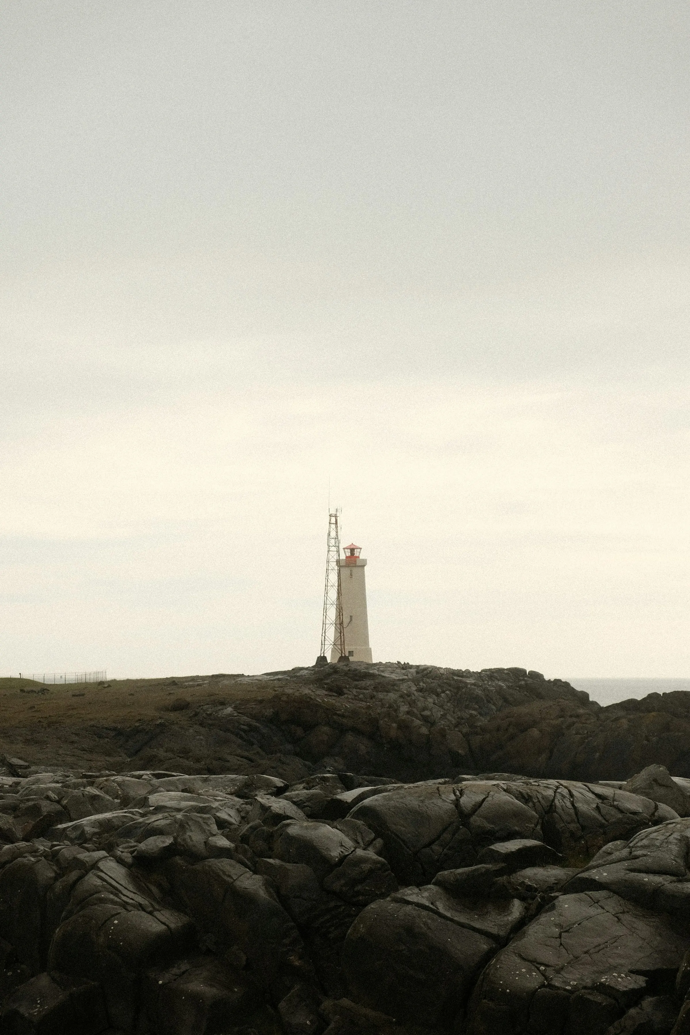 A white lighthouse with a red lantern, a slender mast beside it, standing above dark, fissured rock on the Icelandic coast under a pale overcast sky.