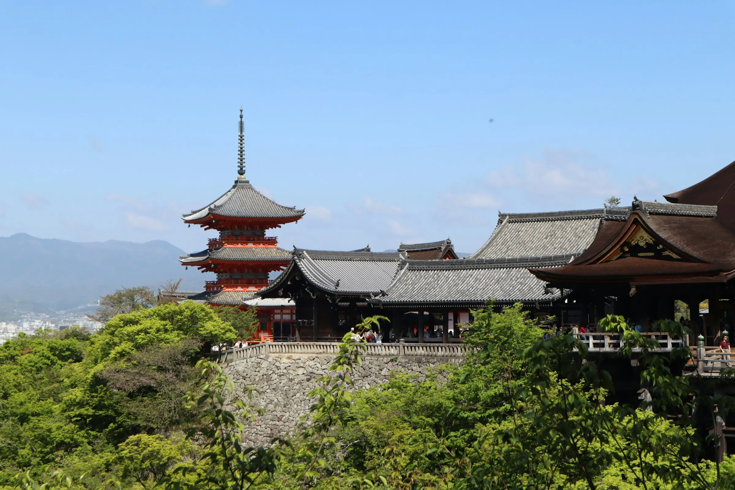 The vermilion pagoda and tiled halls of Kiyomizu-dera above fresh green foliage, with the city of Kyoto and its hills beyond.