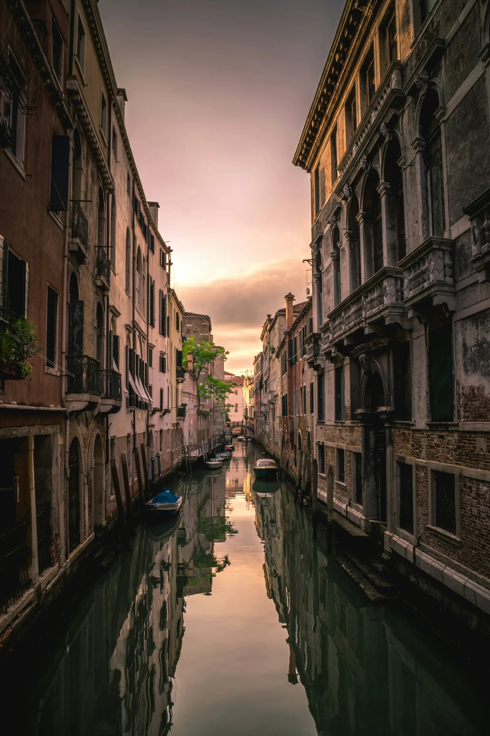 A narrow Venetian canal at dusk, its tall façades and a distant bridge mirrored in still water beneath a rose-coloured sky.