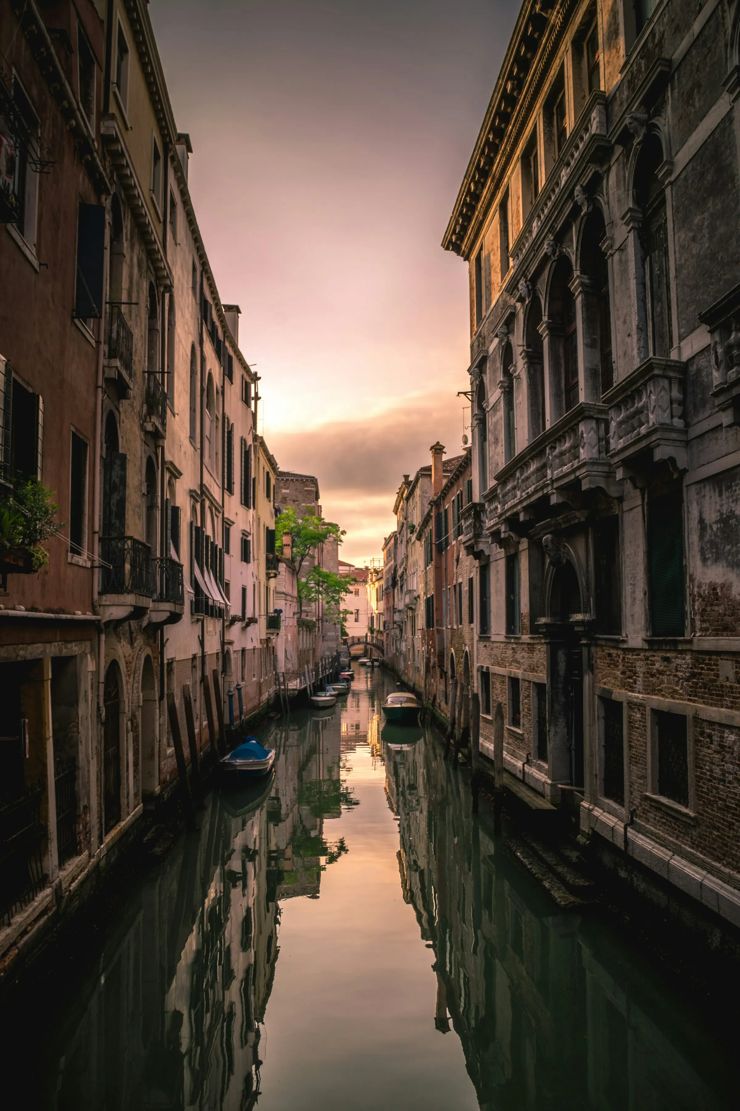 A narrow Venetian canal at dusk, its tall façades and a distant bridge mirrored in still water beneath a rose-coloured sky.